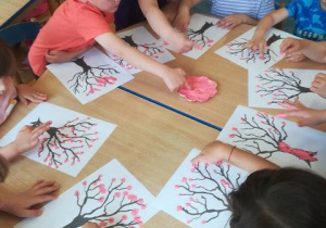 Grupa dzieci przy stoliku stempluje palcem umoczonym w różowej farbie kwitnące drzewo.
